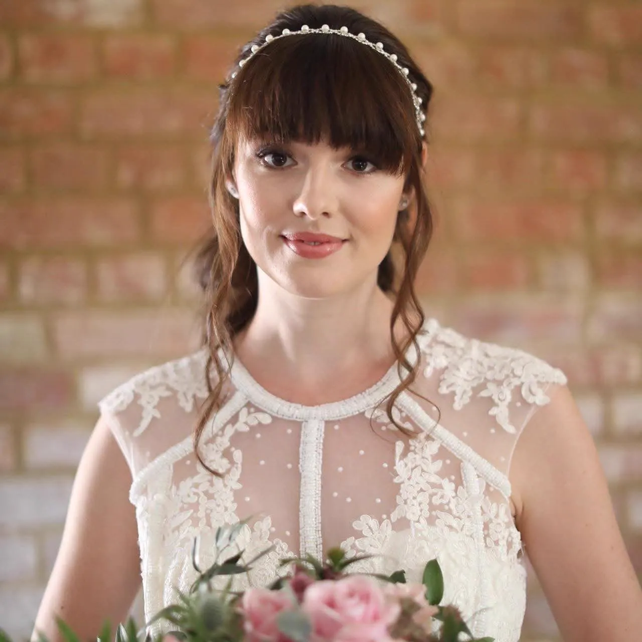 Bride with a pearl headband and soft fringe curls against a brick wall by Be Bridal
