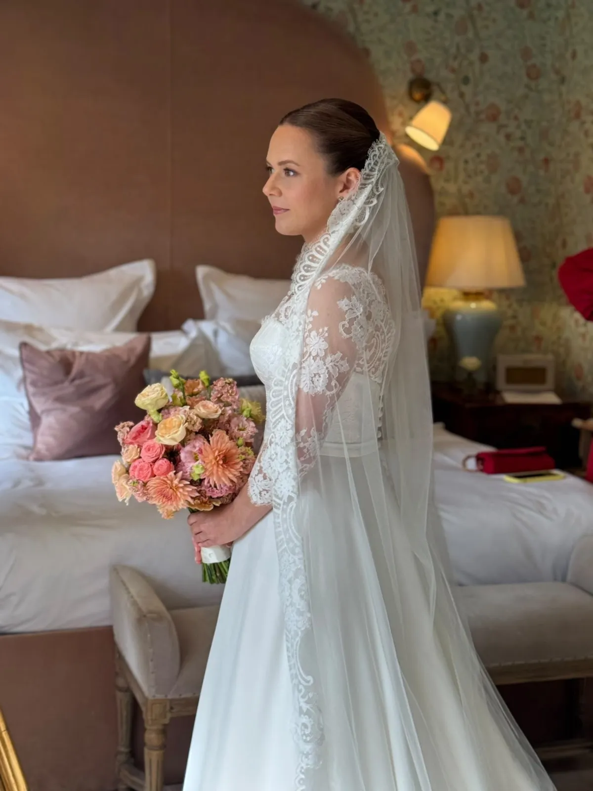Bride in a lace veil with a sleek low bun and peach bouquet by Be Bridal