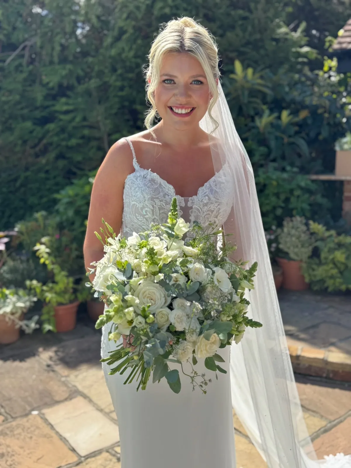 Smiling bride in an embroidered wedding dress holding a bouquet on a stone patio by Be Bridal