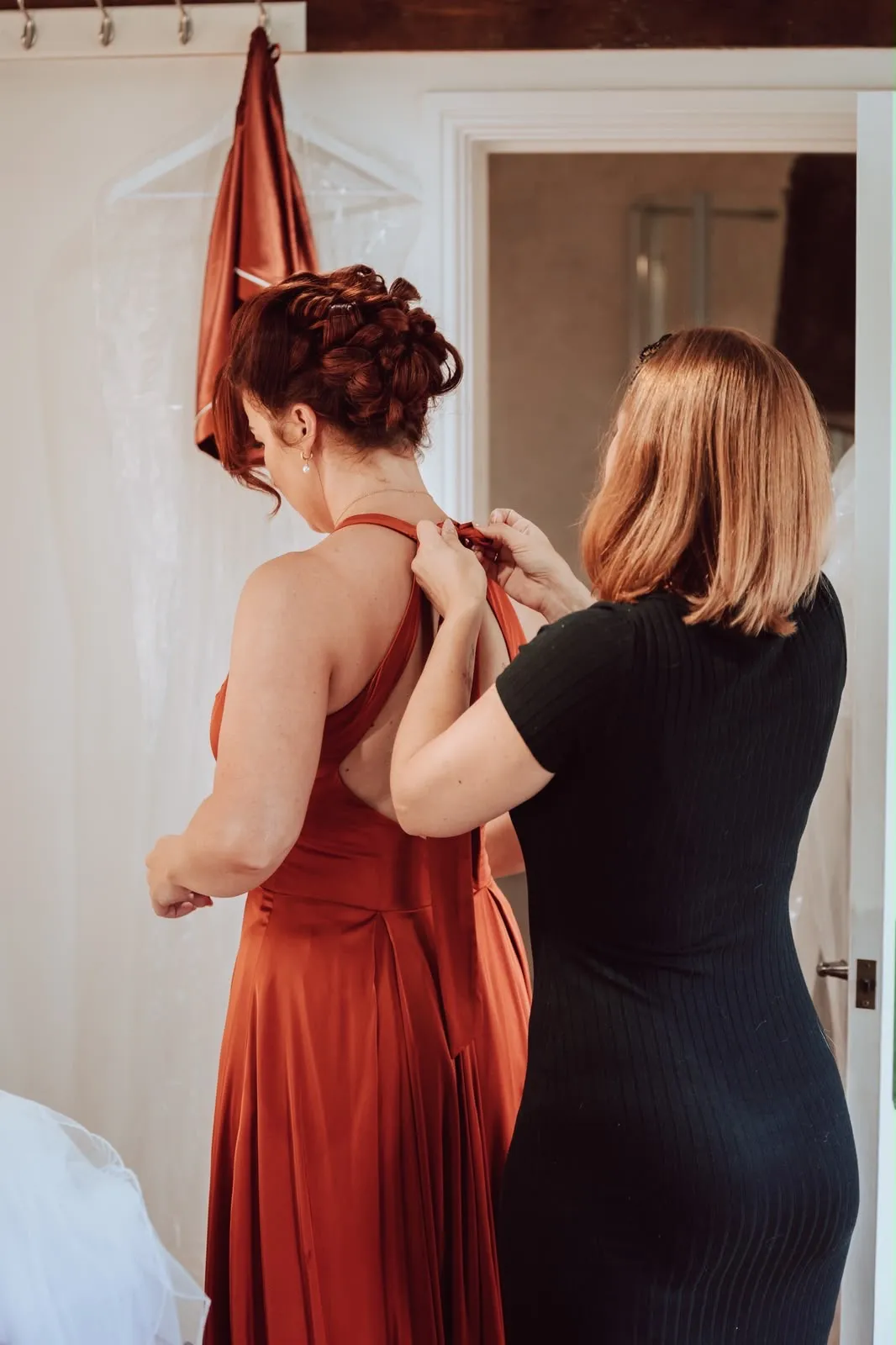 Artist fastening a bridesmaid's rust satin dress after her updo by Be Bridal