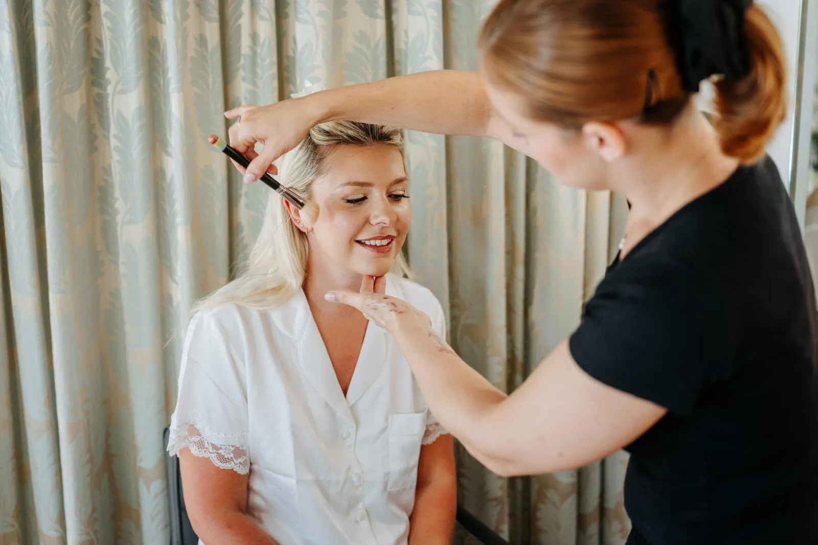 Makeup artist sweeping blush onto a smiling blonde bride by Be Bridal