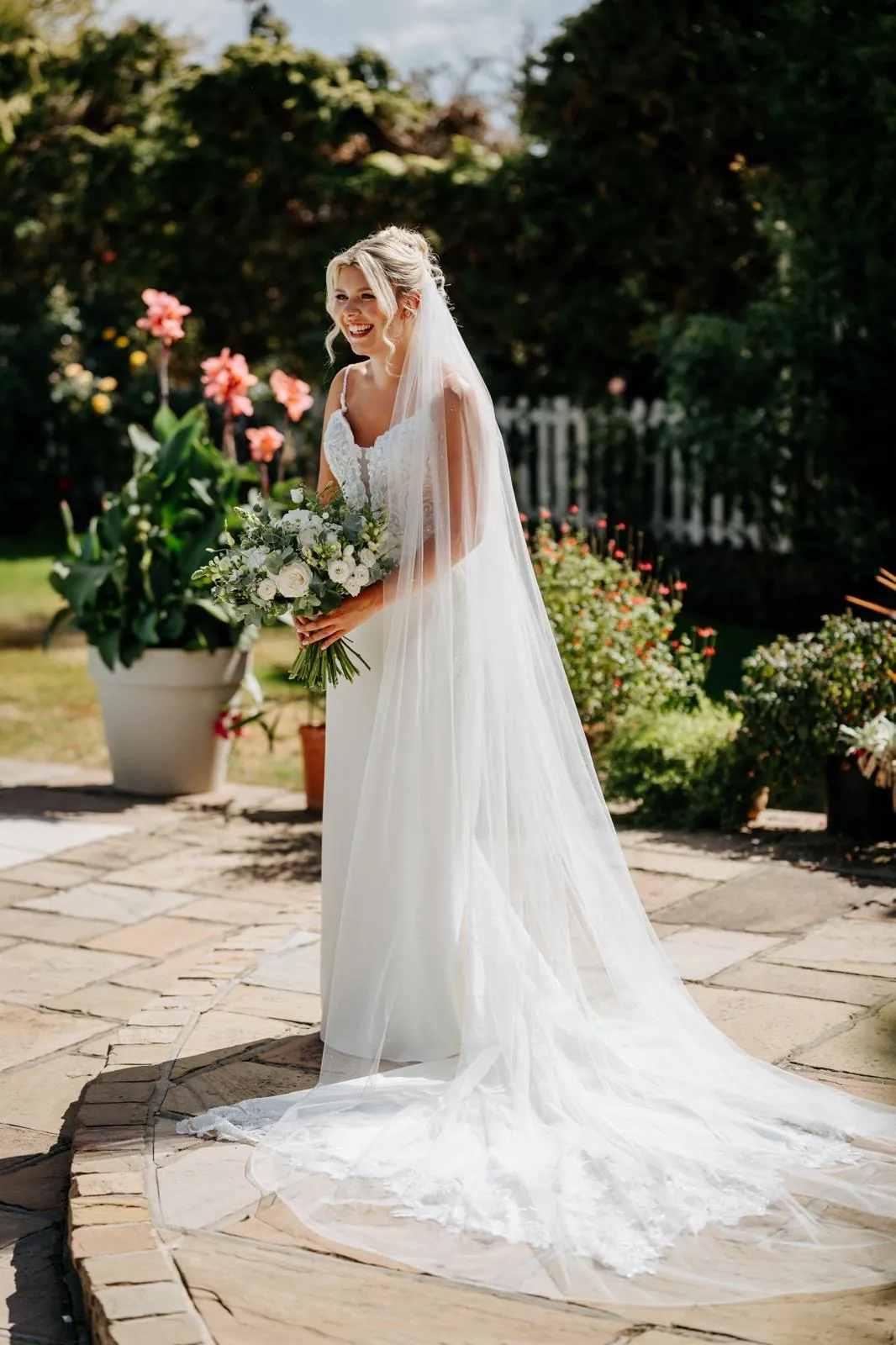 Laughing bride in a lace gown holding a white rose and eucalyptus bouquet on a sunlit stone patio, her cathedral veil trailing across the flagstones by Be Bridal