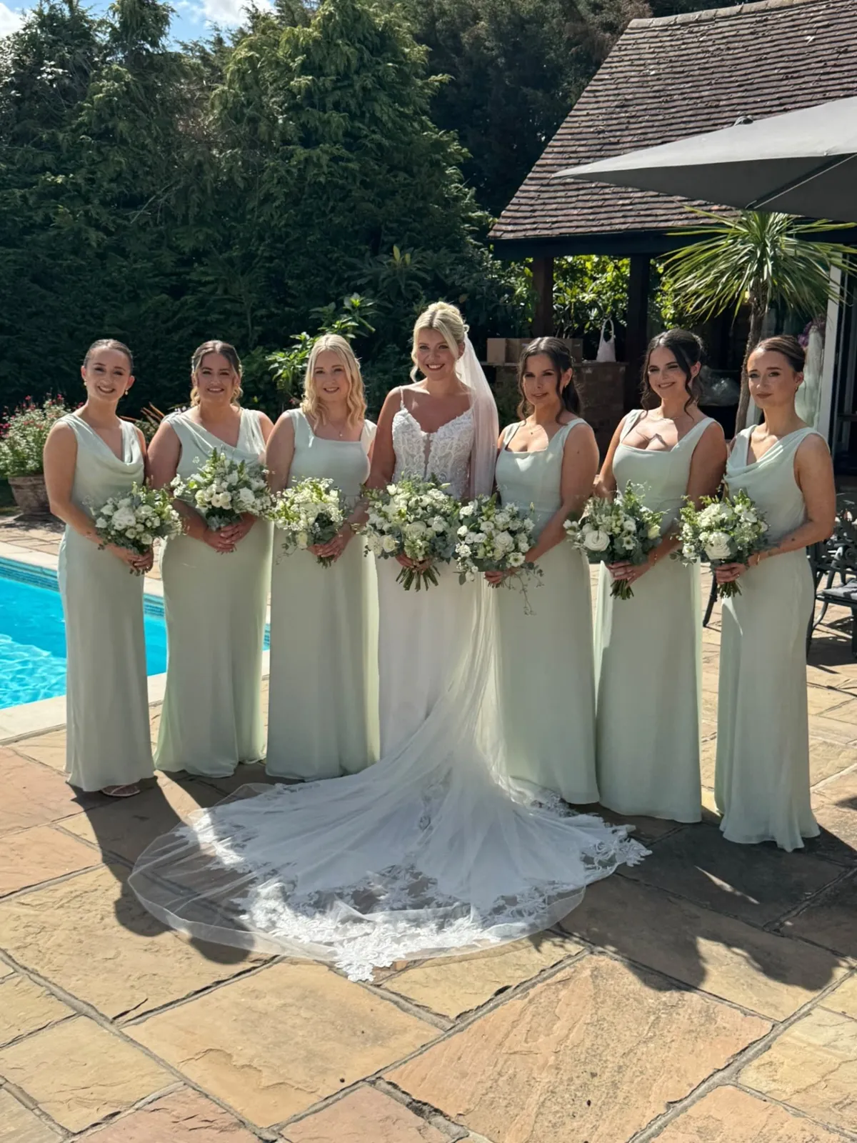 Bride and bridesmaids in sage gowns holding white bouquets by the pool by Be Bridal